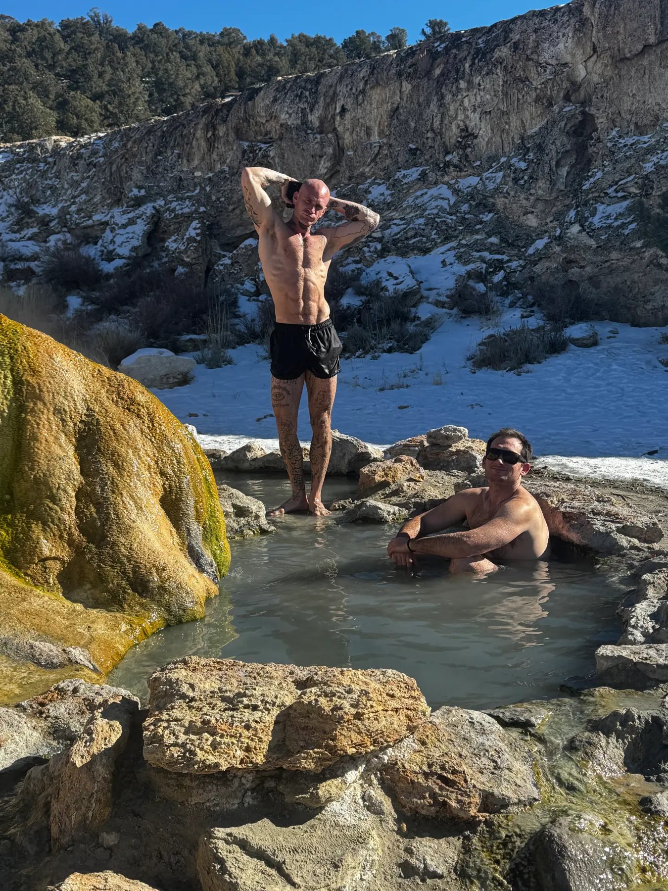 Two men in the roadside hot springs surrounded by snow during a wilderness therapy trip