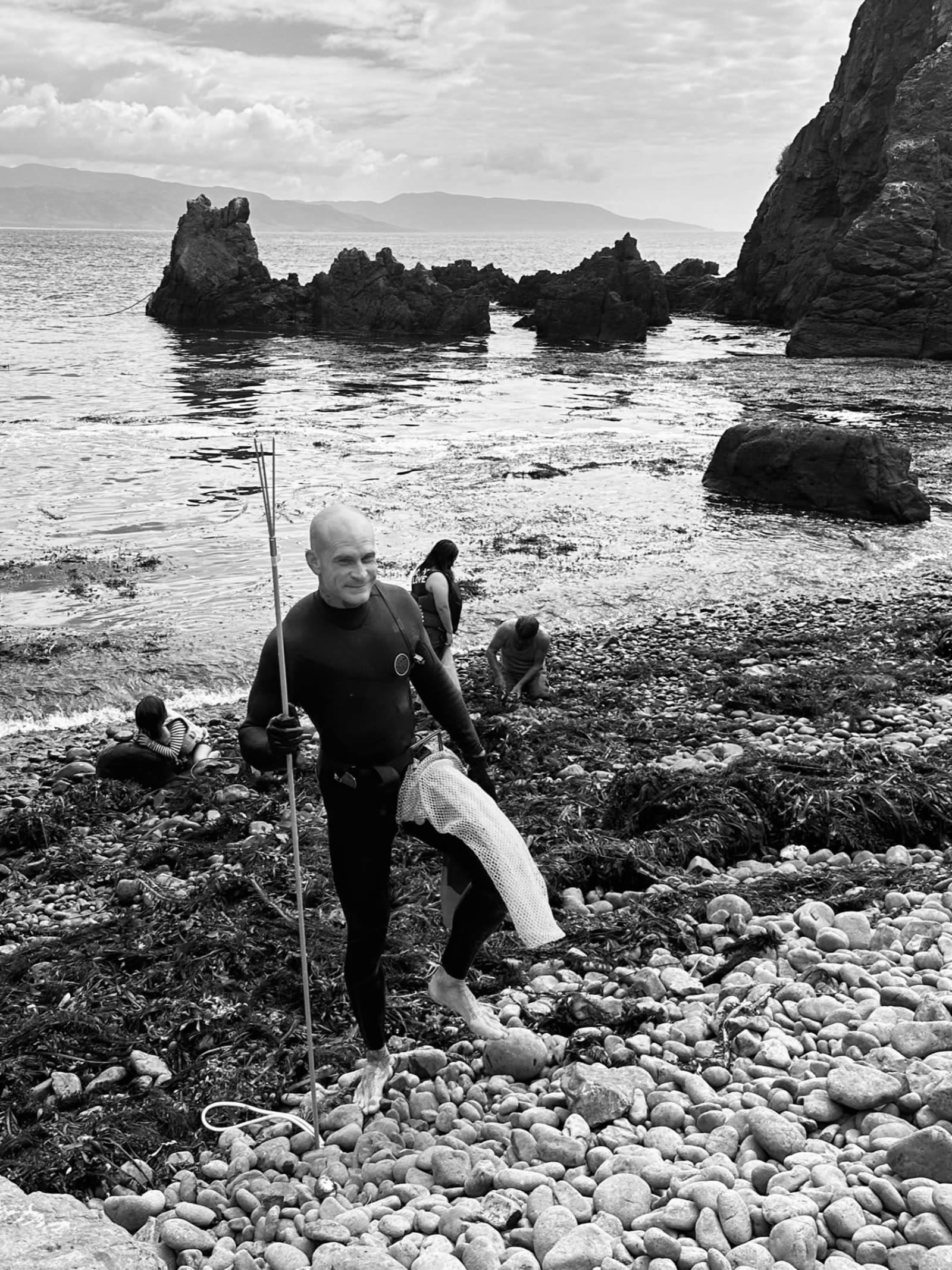 A man walking up the cobble beach in Baja with a spear after spearfishing